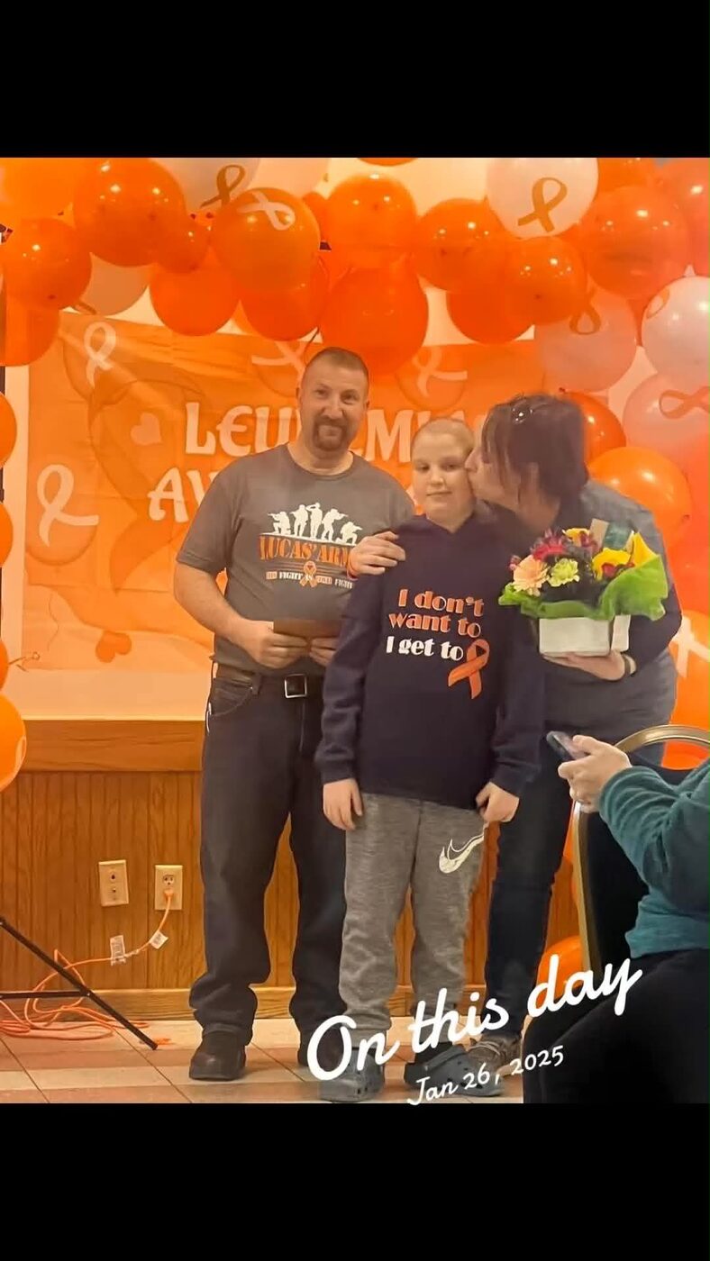 Lucas with his parents on stage under orange balloons at a leukemia awareness night