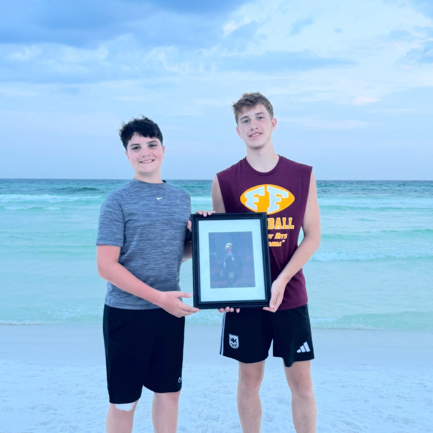 Logan and Andrew standing on the beach holding a framed Forever 12 photograph of their brother Lucas
