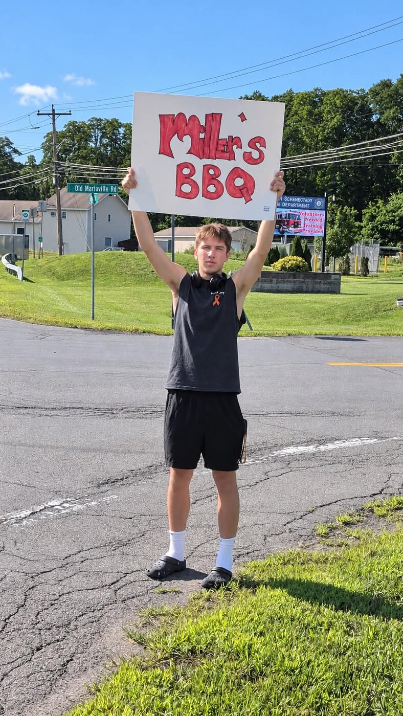 A volunteer holding a hand-lettered Miller's BBQ sign at the roadside
